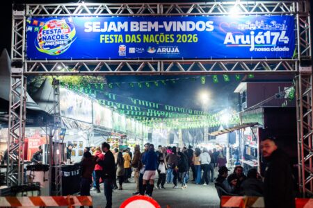 Pórtico de entrada da Festa das Nações 2026 em Arujá, com pessoas circulando entre barracas de comida à noite