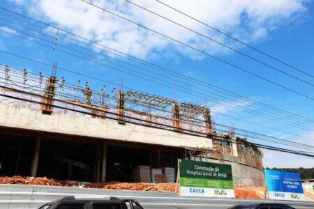 Operários trabalhando na fachada de alvenaria do canteiro de obras do futuro Hospital Municipal de Arujá, sob a supervisão de engenheiros e com maquinário de construção visível ao fundo.